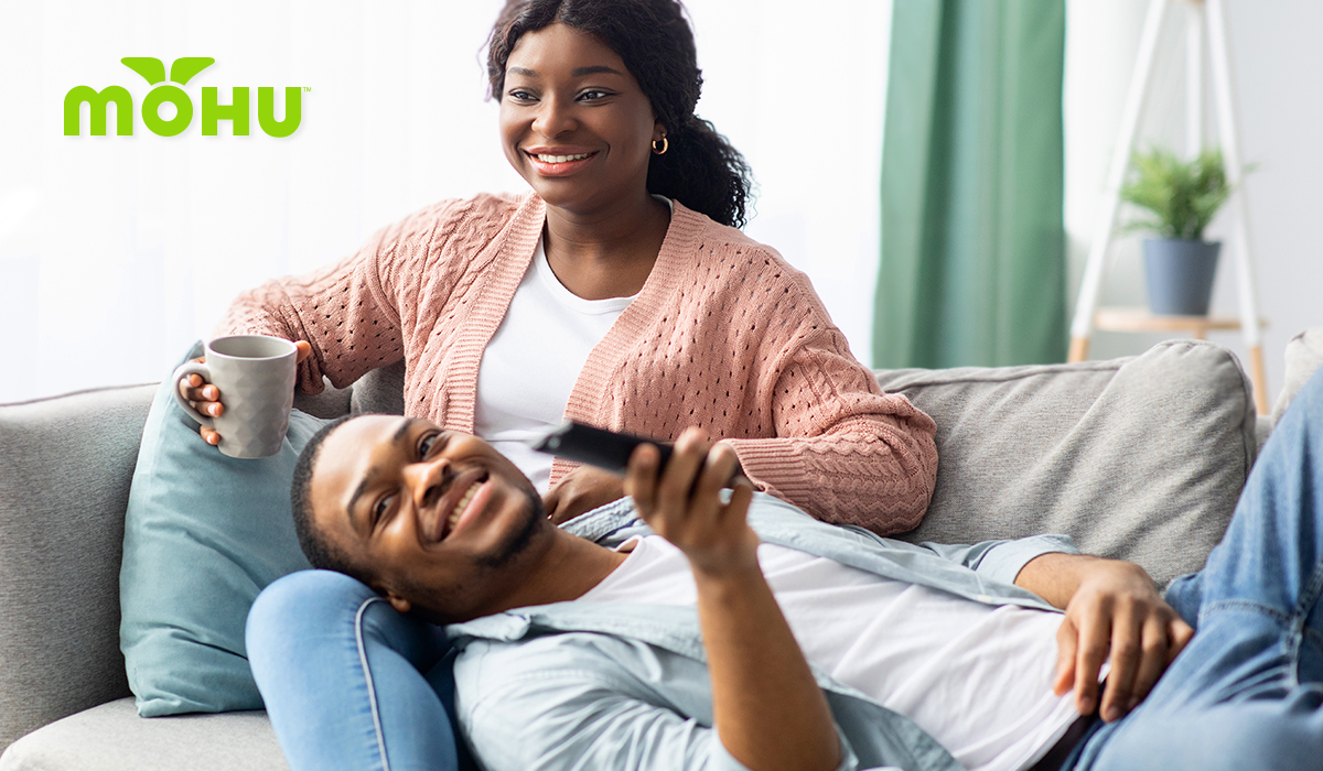 Couple laying on the couch watching Television, flipping through channels with remote in hand. Mohu logo.